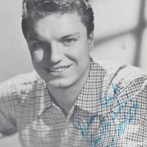May include: Black and white photograph of a smiling man with neatly styled hair. He wears a checkered button-down shirt. The image includes a handwritten blue inscription that reads "Dear Bob, Best wishes, Guy Mitchell."
