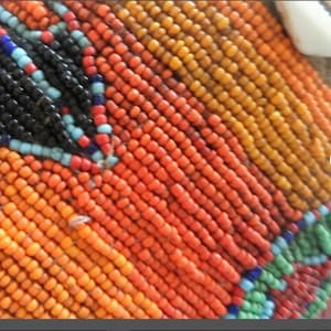 May include: Close-up of a beaded craft with orange, red, yellow, black, and blue beads. The beads are arranged in a geometric pattern. The craft also features white seashells.