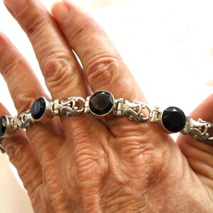 May include: Silver-toned bracelet featuring oval, black gemstones. The bracelet has an ornate design with decorative links. The gemstones are evenly spaced along the bracelet's length, creating a classic and elegant look.