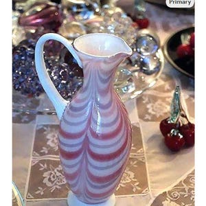 May include: A pink and white striped glass pitcher with a white handle and base. The pitcher has a curved spout and a bulbous body. Decorative glassware.