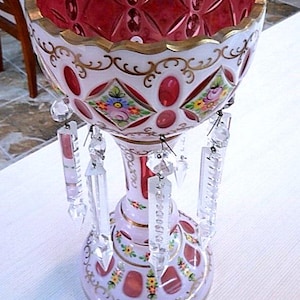 May include: An ornate, vintage glass vase with a ruby red interior and a white exterior adorned with gold trim and floral designs. The vase features hanging crystal accents and is a decorative piece.