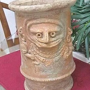 May include: A cylindrical, terracotta-colored vase with a detailed, carved face design. The vase features a wide rim and a textured surface, with additional carved figures on the sides. The vase is displayed on a red surface.