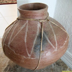 May include: A large, round, earthenware pot with a reddish-brown hue. The pot features white painted lines and is bound with a natural fiber. The pot has a wide opening and a slightly textured surface. A decorative piece.