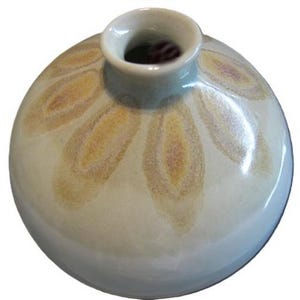May include: A small, round ceramic vase with a wide body and a narrow neck. The vase is primarily off-white with a pattern of stylized, petal-like shapes in shades of yellow and brown. The rim is a light blue-gray color.
