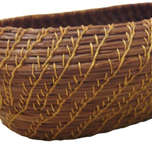 May include: A handwoven, oval-shaped basket with a natural brown hue. The basket features intricate, decorative stitching in a contrasting golden-yellow thread, creating a textured, patterned surface. The basket's design suggests it could be used for storage or display.