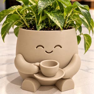 May include: A matte beige ceramic planter shaped like a character with arms holding a small cup and saucer. The planter features a simple carved smiling face and contains a variegated green and yellow pothos plant with heart-shaped leaves.