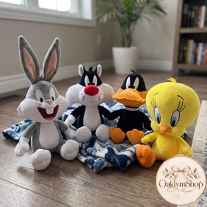 Op de afbeelding: Een set van vier pluche speelgoedfiguren met Bugs Bunny, Sylvester, Daffy Duck en Tweety zittend op een blauw-witte deken met patroon. Elk personage is uitgevoerd in de klassieke kleuren, waaronder grijs, zwart, wit, geel en oranje.