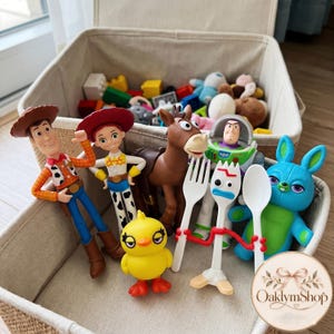 May include: A collection of plastic character figures including a cowboy, a cowgirl, a brown horse, a space ranger, a blue rabbit, a yellow chick, and a white spork character, all arranged inside a beige fabric storage bin with other toys.