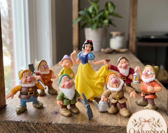 May include: A set of eight plastic figurines featuring Snow White in a blue and yellow dress surrounded by seven dwarfs in various colored outfits, including brown, green, and red, with accessories like a pickaxe, lantern, and watering can.
