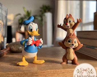 Può includere: Una statuetta di Paperino con camicia da marinaio blu e papillon rosso, accanto a una coppia di statuette di Cip e Ciop impilate. I personaggi sono realizzati in stile cartoon con colori vivaci, caratterizzati dal cappello di Paperino e dal pelo marrone.