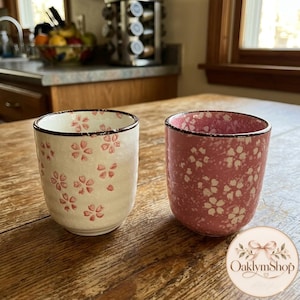 Puede incluir: Dos tazas de cerámica de forma cilíndrica con un borde marrón oscuro. La taza de la izquierda es de color blanco roto con un patrón de flores de cerezo rosas. La taza de la derecha es rosa con un patrón de flores de cerezo blancas y textura moteada.