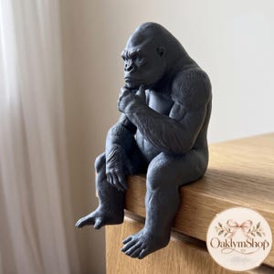 Op de afbeelding: Een matzwart gorillabeeldje in een zittende, peinzende houding met één hand bij de kin. De gorilla heeft een gedetailleerde vachtstructuur en spierdefinitie, ontworpen om op de rand van een oppervlak te zitten. De tekst OaklymShop is zichtbaar.