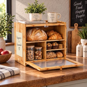 May include: A bamboo bread box with a clear, ribbed drop-down door and a side panel featuring the text "Happiness is Homemade". The interior contains a large loaf of bread, two glass jars filled with nuts and grains, muffins, and rolls.