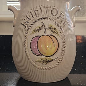 May include: A cream-coloured ceramic Rumtopf jar with a matching lid and two side handles. The exterior features vertical ribbing and a central embossed oval medallion depicting a peach and a plum with leaves, surrounded by a rope-like border design.