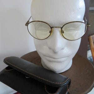 May include: Oval-framed eyeglasses with a gold-tone metal frame and yellow-tinted lenses rest on a white mannequin head. A black leather eyeglass case with brown stitching sits below the glasses. The case is closed.