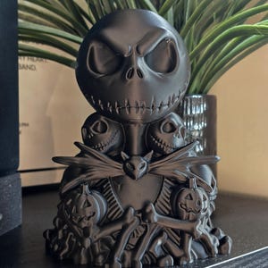 May include: A matte black 3D printed figurine featuring a large central skull character with hollow eyes and a stitched mouth, surrounded by smaller skull heads, two carved jack-o'-lantern pumpkins, and a bat-shaped bowtie at the centre.