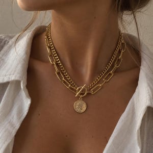 May include: A layered gold-tone necklace featuring a delicate curb chain and a thicker elongated oval link chain. The piece is secured with a front-facing toggle clasp that holds a circular pendant embossed with a profile portrait of a figure.