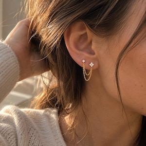 May include: A gold double chain earring featuring two stud components, one with a single round clear stone and the other with a four-stone flower-shaped cluster, connected by two delicate gold chains that drape below the earlobe.