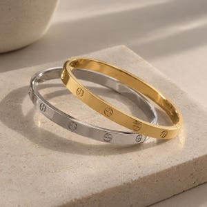 Può includere: Due bracciali rigidi incernierati, uno in tonalità oro lucido e uno in tonalità argento lucido. Ogni bracciale presenta una superficie liscia e piatta decorata con motivi circolari incisi e distanziati uniformemente contenenti un simbolo a croce.