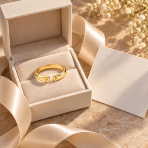 Puede incluir: Un anillo abierto de tono dorado con las letras M y K grabadas en los extremos, presentado dentro de un joyero beige con una tarjeta en blanco a juego y una cinta de satén. El anillo tiene un acabado liso y pulido con una banda ajustable.