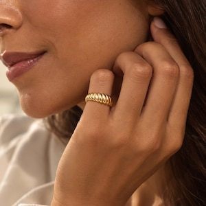 May include: A gold-toned ring featuring a thick, rounded band with a repeating diagonal ribbed or twisted texture. The metal surface has a polished, reflective finish that catches the light, creating a series of smooth, curved ridges along the band.