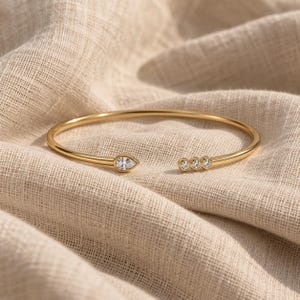 May include: A gold-toned open cuff bracelet featuring a single pear-shaped clear stone at one end and three small, round clear stones set in a row at the opposite end. The metal band is smooth and polished, creating a continuous circular shape.