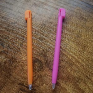 Puede incluir: Dos lápices ópticos de plástico, uno de color naranja brillante y otro de color rosa vibrante, cada uno con un cuerpo cilíndrico delgado, un pequeño clip rectangular en el extremo superior y una punta redondeada transparente para pantallas táctiles.