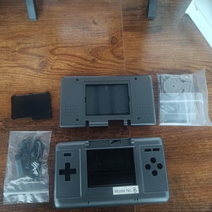 Puede incluir: Un kit de carcasa de repuesto para consola de juegos portátil en color gris oscuro que incluye una carcasa frontal con una etiqueta Model No. B, una carcasa trasera, una tapa de batería negra y dos bolsas de plástico con botones negros.