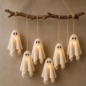 Op de afbeelding: Een wandhanger bestaande uit een natuurlijke houten tak opgehangen aan touw, waaraan zes gehaakte gebroken witte spookfiguren hangen. Elk spookje bevat een klein, warm gloeiend lampje en heeft twee kleine zwarte ronde ogen.