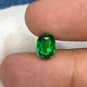 May include: An oval-cut gemstone with a vibrant, saturated green color and a faceted surface that reflects light. The stone is translucent, displaying deep emerald tones and clear internal clarity, showcasing a polished finish and precise geometric cuts.
