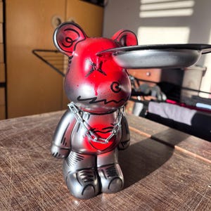 以下が含まれることがあります： メタリックシルバー仕上げのクマ型フィギュア。頭部と胸部に赤いスプレー塗装のようなエフェクトが施されています。黒いX型の目、シルバーのチェーンネックレス、頭部に取り付けられた黒い円形のトレイが特徴です。