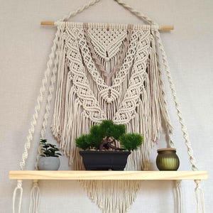 Peut inclure: Une étagère en bois fixée au mur et suspendue par une corde en macramé crème. L'étagère présente une toile de fond en macramé avec des motifs noués complexes et de longues franges. Une petite plante verte et un pot en verre sont posés dessus.