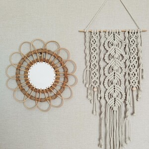 Peut inclure: Un miroir rond avec un cadre en rotin marron clair au design en forme de pétales et une suspension en macramé faite de corde de coton blanc cassé, accrochée à une tige en bois avec des motifs noués complexes et des franges pendantes.