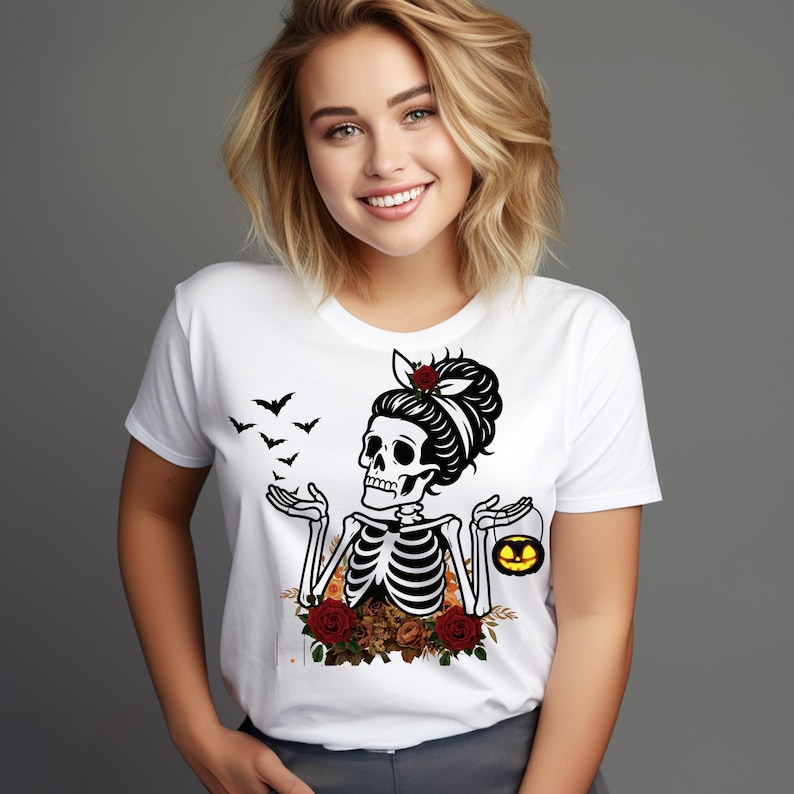 May include: A white t-shirt featuring a graphic print of a skeleton with a hair bun, red roses, and bats. The skeleton holds a small glowing jack-o'-lantern bucket. The design includes floral elements at the base and a white background.