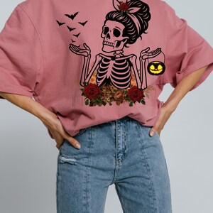 May include: A dusty rose t-shirt featuring a black graphic of a skeleton with a hair bun, holding a small glowing jack-o'-lantern. The skeleton is surrounded by red roses and orange flowers, with a flock of black bats flying above one hand.