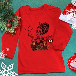 May include: A long-sleeve red shirt featuring a black graphic of a skeleton with a messy bun hairstyle, holding a small yellow jack-o'-lantern bucket. The skeleton is surrounded by red roses and a flock of black bats flying toward its open hands.