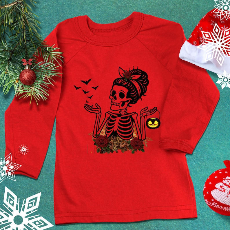May include: A long-sleeve red shirt featuring a black graphic of a skeleton with a messy bun hairstyle, holding a small yellow jack-o'-lantern bucket. The skeleton is surrounded by red roses and a flock of black bats flying toward its open hands.