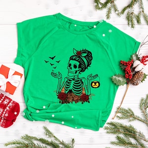 May include: A bright green short-sleeve t-shirt featuring a black graphic of a skeleton with a hair bun, red roses, and bats. The skeleton holds a small orange jack-o'-lantern bucket in one hand and has its other hand raised with bats flying.