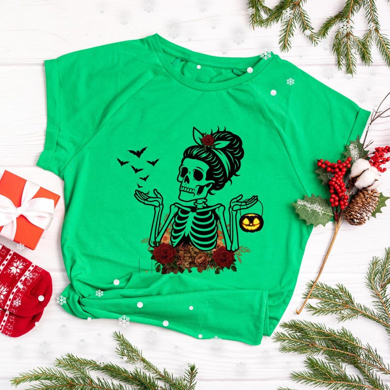 May include: A bright green short-sleeve t-shirt featuring a black graphic of a skeleton with a hair bun, red roses, and bats. The skeleton holds a small orange jack-o'-lantern bucket in one hand and has its other hand raised with bats flying.