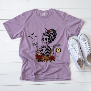 May include: A light purple short-sleeve t-shirt featuring a graphic of a skeleton with a hair bow, surrounded by red roses and autumn leaves. The skeleton holds a small glowing jack-o'-lantern, with black bats flying nearby.