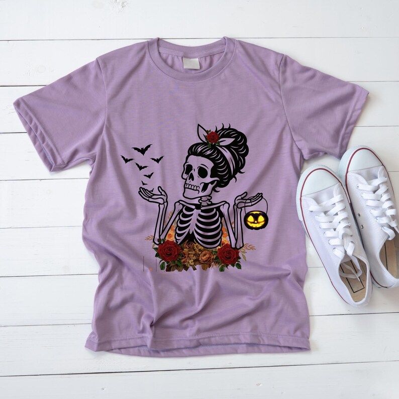 May include: A light purple short-sleeve t-shirt featuring a graphic of a skeleton with a hair bow, surrounded by red roses and autumn leaves. The skeleton holds a small glowing jack-o'-lantern, with black bats flying nearby.