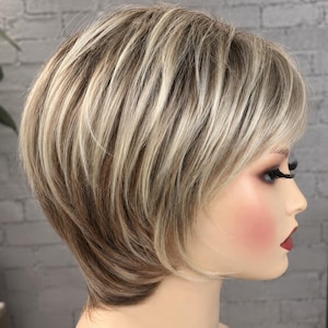 May include: A short, layered wig featuring a blend of light blonde and medium brown highlights. The hair is styled with a side-swept fringe and textured layers that frame the face, showcasing a multi-tonal color palette with darker roots.