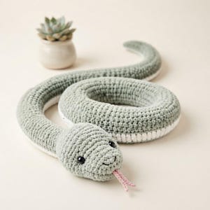 May include: A sage green and white crocheted snake toy with a coiled body, black bead eyes, and a small pink forked tongue. The snake is crafted with a textured yarn stitch pattern, featuring a light green top and a solid white underside.