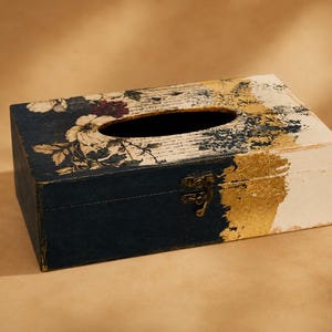 May include: A rectangular wooden tissue box cover featuring a dark navy blue base, a white section, and gold leaf accents. The top surface displays a floral pattern with white and dark red flowers layered over a background of printed script text.