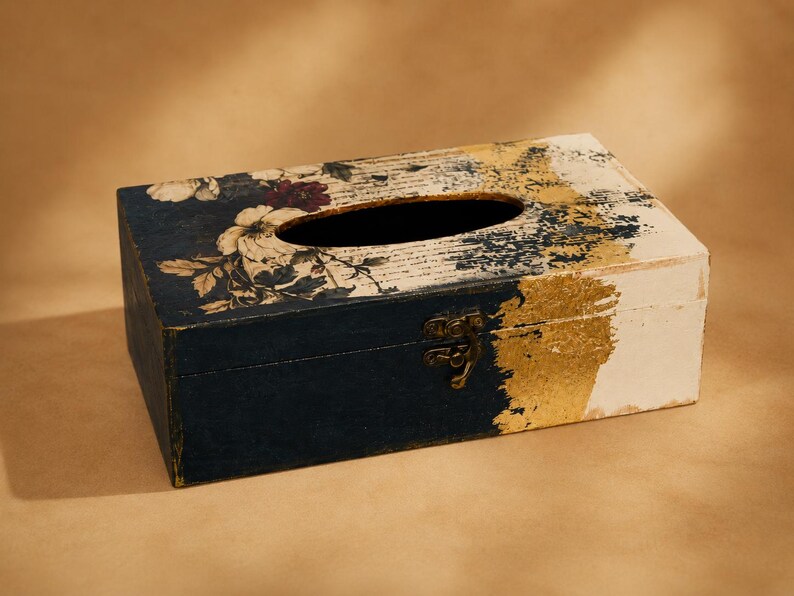 May include: A rectangular wooden tissue box cover featuring a dark navy blue base, a white section, and gold leaf accents. The top surface displays a floral pattern with white and dark red flowers layered over a background of printed script text.