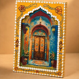 May include: A rectangular wall hanging featuring a painted scene of a wooden double door set within a blue archway. The frame is painted yellow with white diamond-shaped accents and floral motifs, and includes a small metal hook at the top.