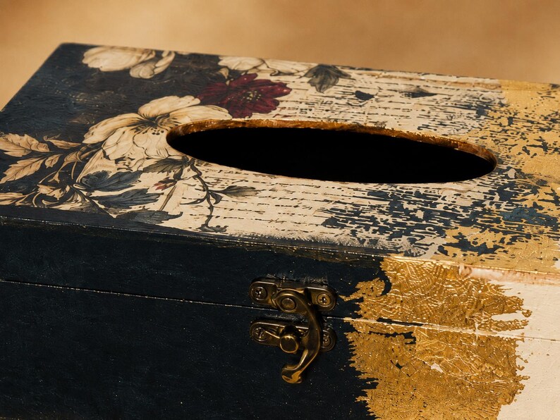 May include: A rectangular wooden tissue box cover featuring a black base with gold leaf accents. The top surface displays a floral pattern with cream and dark red flowers over a background of vintage-style script text and a central oval opening.