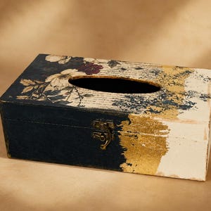 May include: A rectangular tissue box cover featuring a dark navy blue finish on the left side and a cream white finish on the right. The top surface displays a floral pattern with script text, accented by a metallic gold leaf texture in the centre.