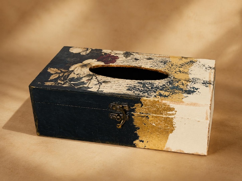 May include: A rectangular tissue box cover featuring a dark navy blue finish on the left side and a cream white finish on the right. The top surface displays a floral pattern with script text, accented by a metallic gold leaf texture in the centre.