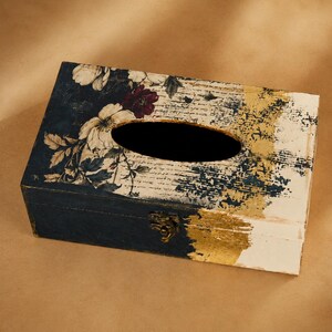 May include: A rectangular wooden tissue box cover featuring a dark blue base with a floral pattern of white and dark red flowers on the left side. The right side displays a distressed white finish with gold leaf accents and a metal latch.